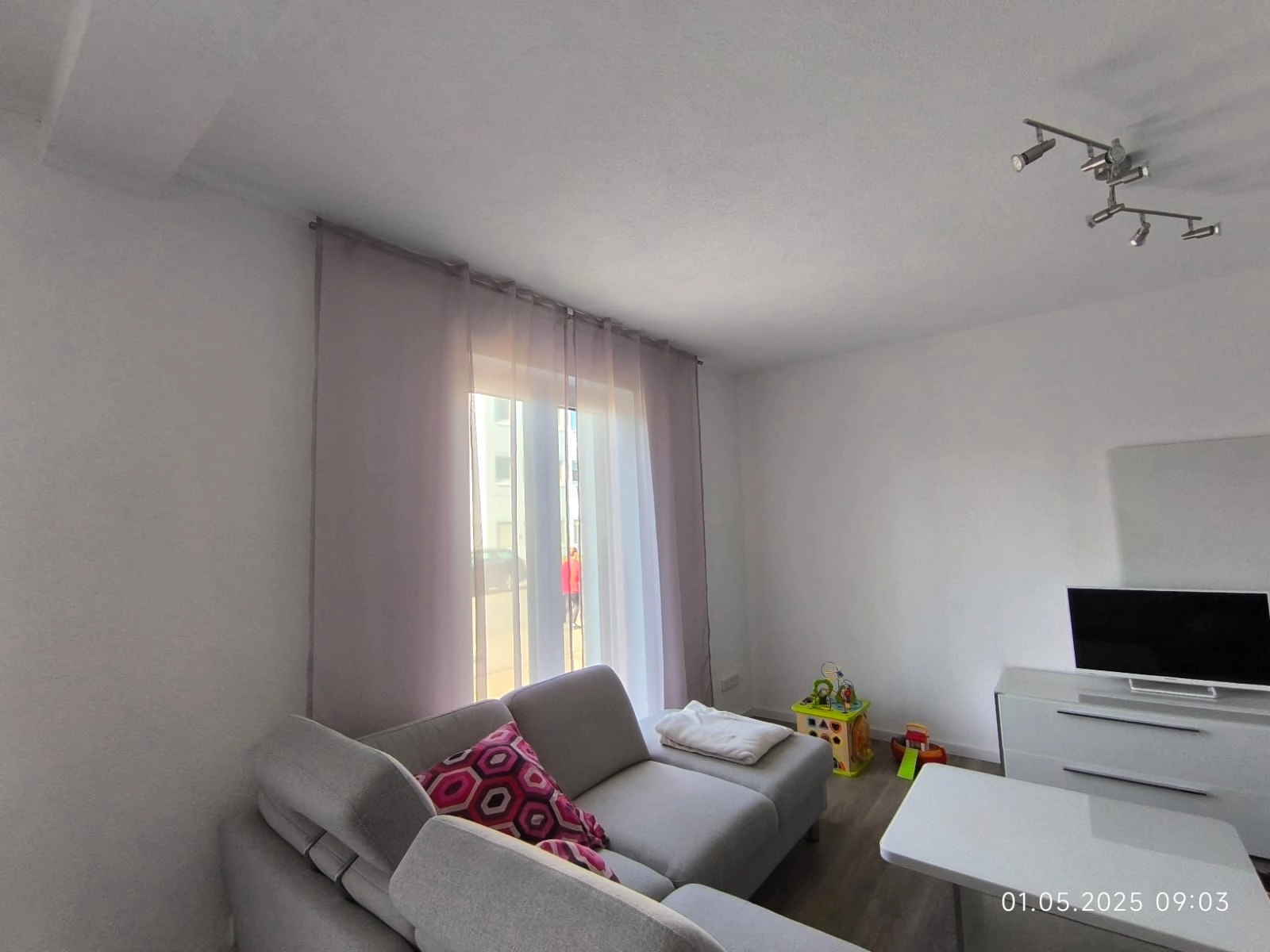 Wohnzimmer mit grauem Sofa, bunten Kissen, Couchtisch, Fernseher und Kinderspielzeug neben einem Fenster.