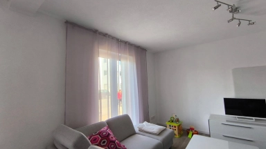 Wohnzimmer mit grauem Sofa, bunten Kissen, Couchtisch, Fernseher und Kinderspielzeug neben einem Fenster.