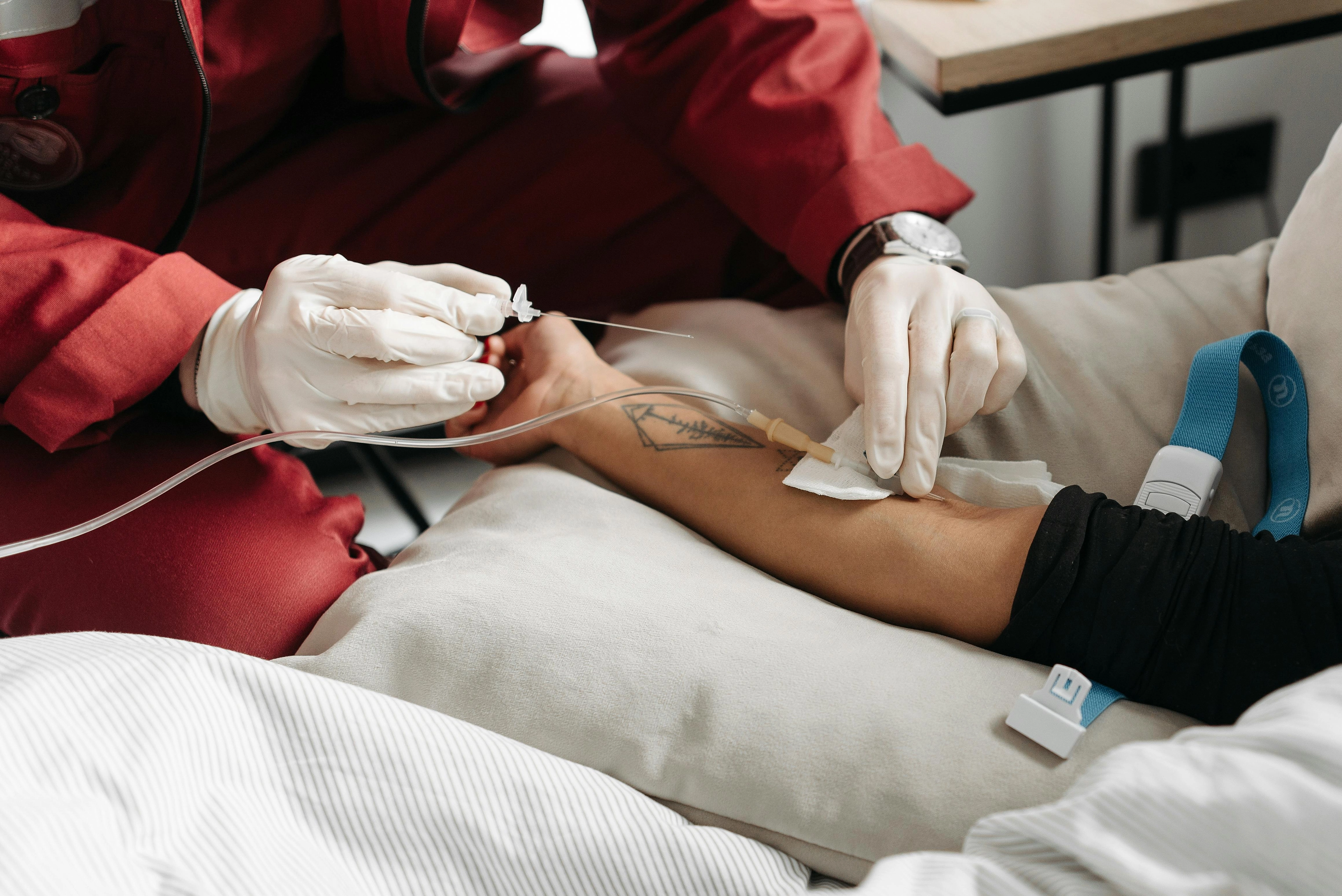 Nahaufnahme: Handschuhe tragende medizinische Fachkraft setzt eine intravenöse Kanüle in den Unterarm einer liegenden Person; Tourniquet und Verbandtupfer sichtbar.