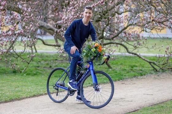 Person auf einem Fahrrad auf einem Parkweg, am Lenker ein großer Blumenstrauß, blühende Bäume im Hintergrund.