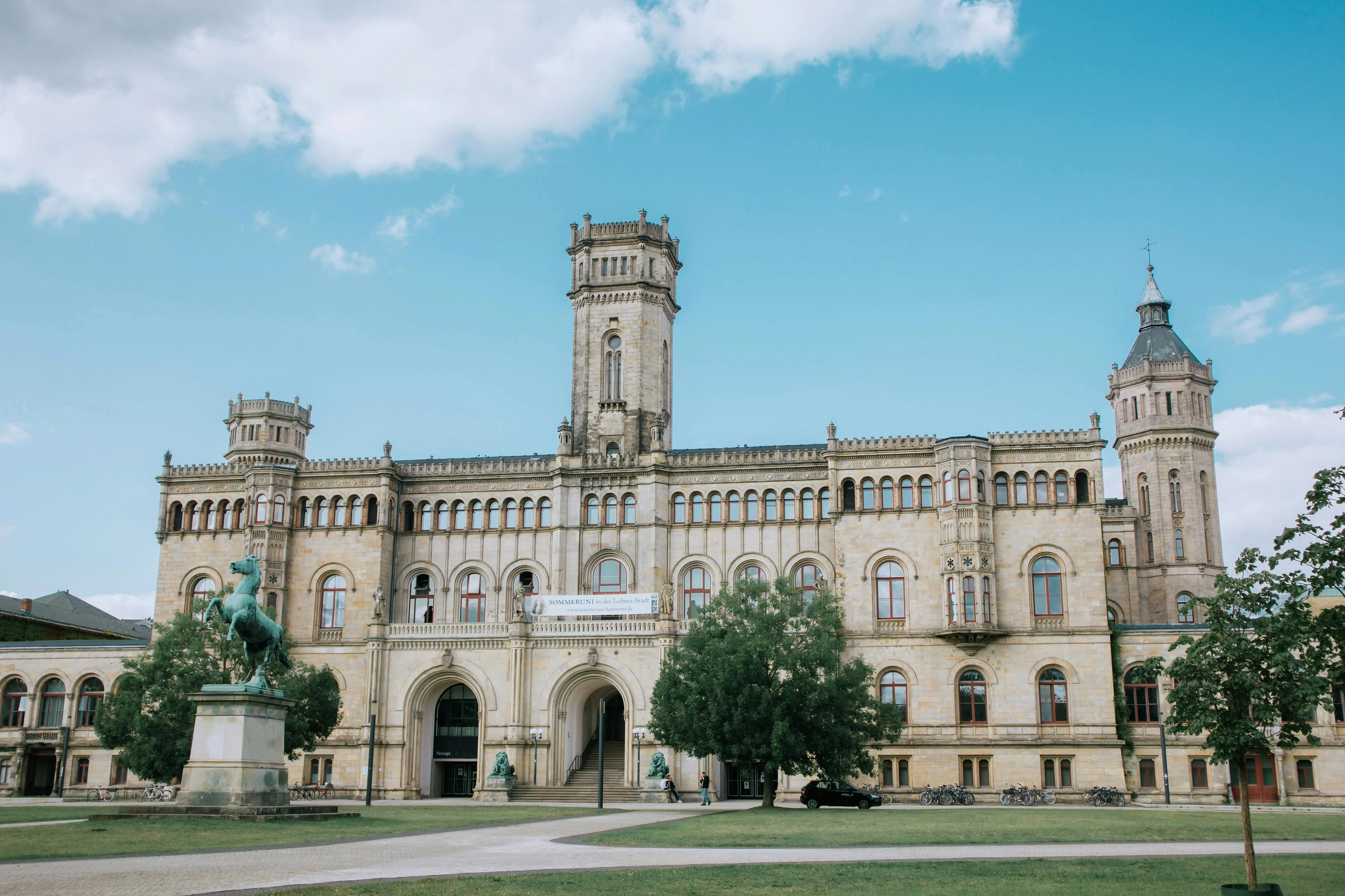Historisches Sandsteingebäude mit zentralem Turm, Bogenportal, einer Reiterstatue im Vordergrund und Grünflächen.