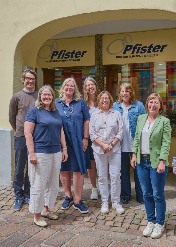 Sieben lächelnde Erwachsene stehen vor dem Schaufenster eines Optikergeschäfts mit Pfister-Logo.