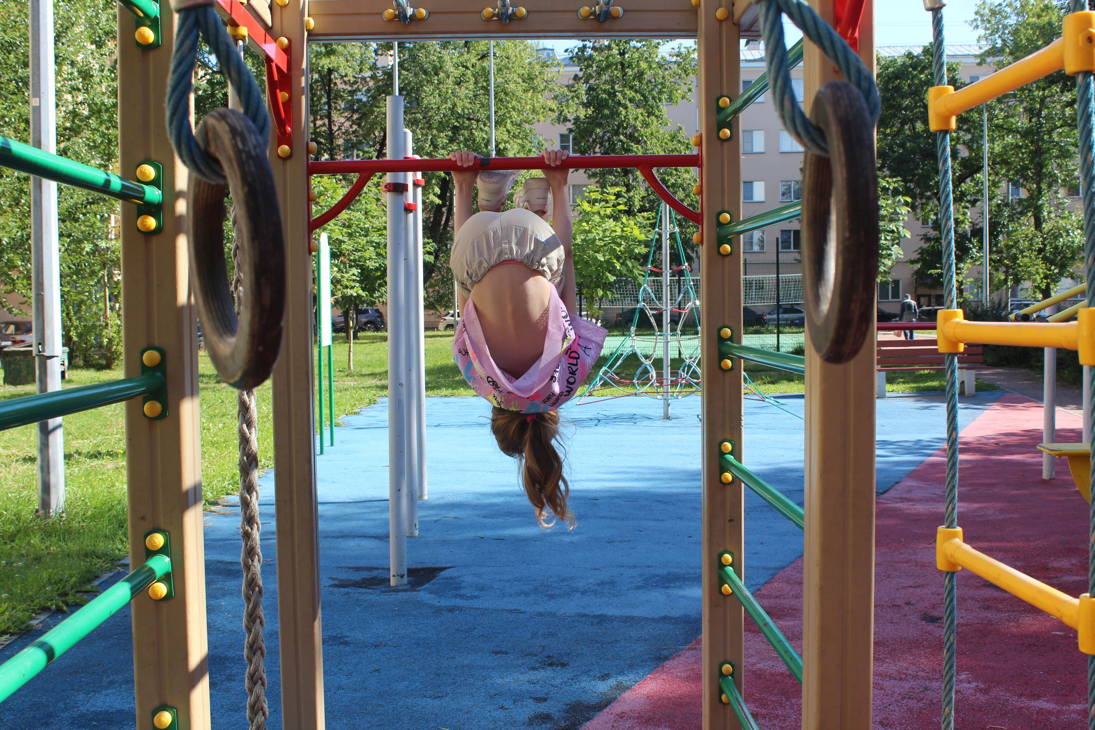 Ein Kind hängt kopfüber an einer Kletterstange auf einem Spielplatz, umgeben von Spielgeräten und Gummibelag.