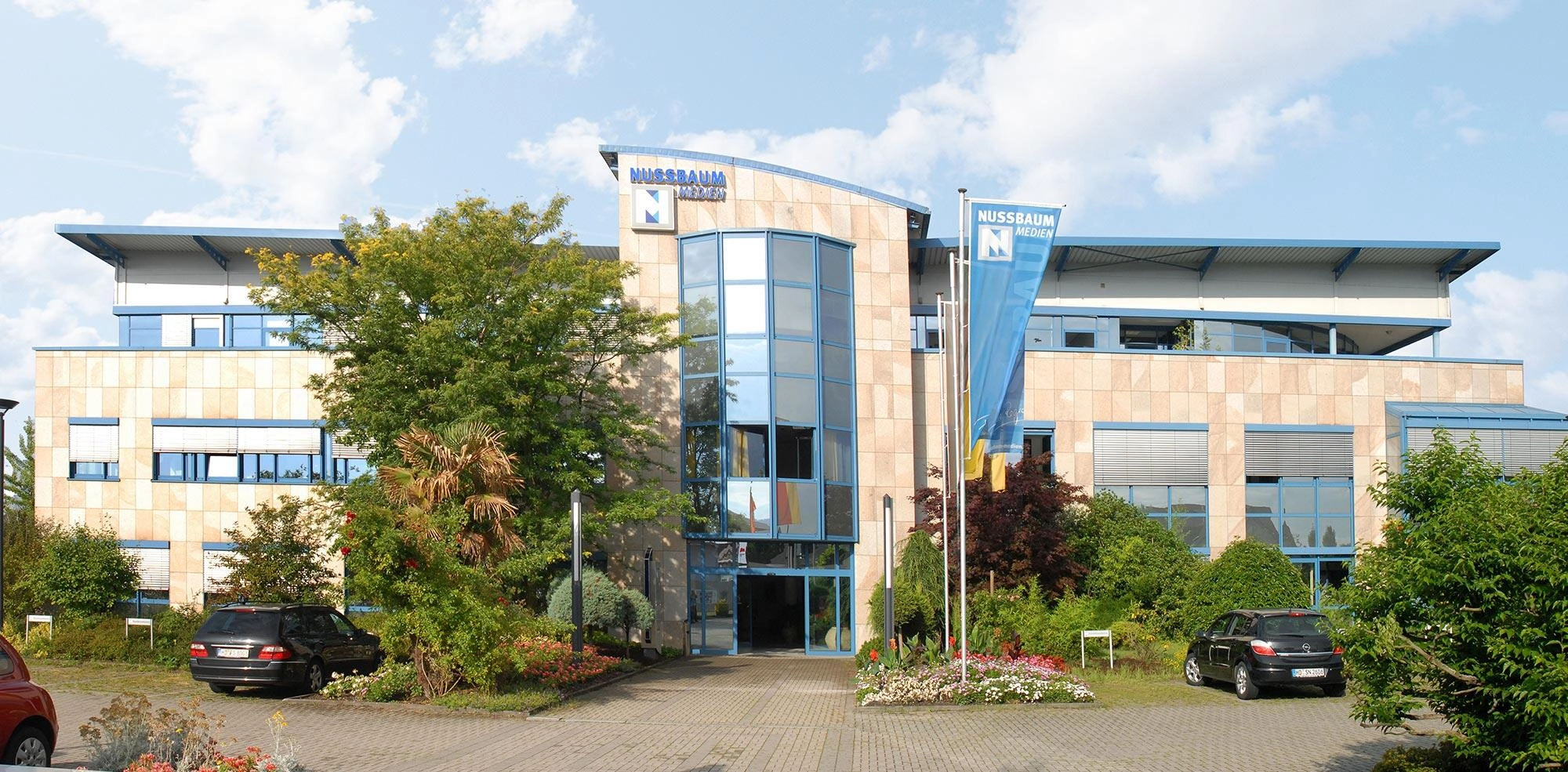Modernes Bürogebäude mit Glasfront, Fahnenmast, Bäumen und Parkplätzen im Vordergrund.