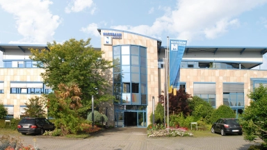 Modernes Bürogebäude mit Glasfront, Fahnenmast, Bäumen und Parkplätzen im Vordergrund.
