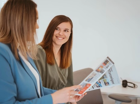 Zwei Frauen sitzen im Gespräch; eine zeigt eine Broschüre, die andere lächelt.