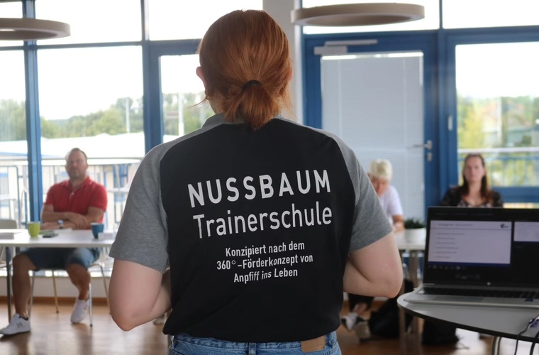 Rückenansicht einer Person mit T-Shirt „NUSSBAUM Trainerschule“ steht vor einer kleinen Gruppe in einem Seminarraum.