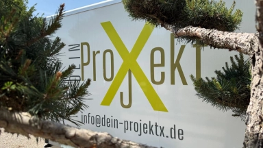 Werbeschild mit dem Logo "ProXekt" und einer E‑Mail‑Adresse, im Vordergrund Kiefernzweige.