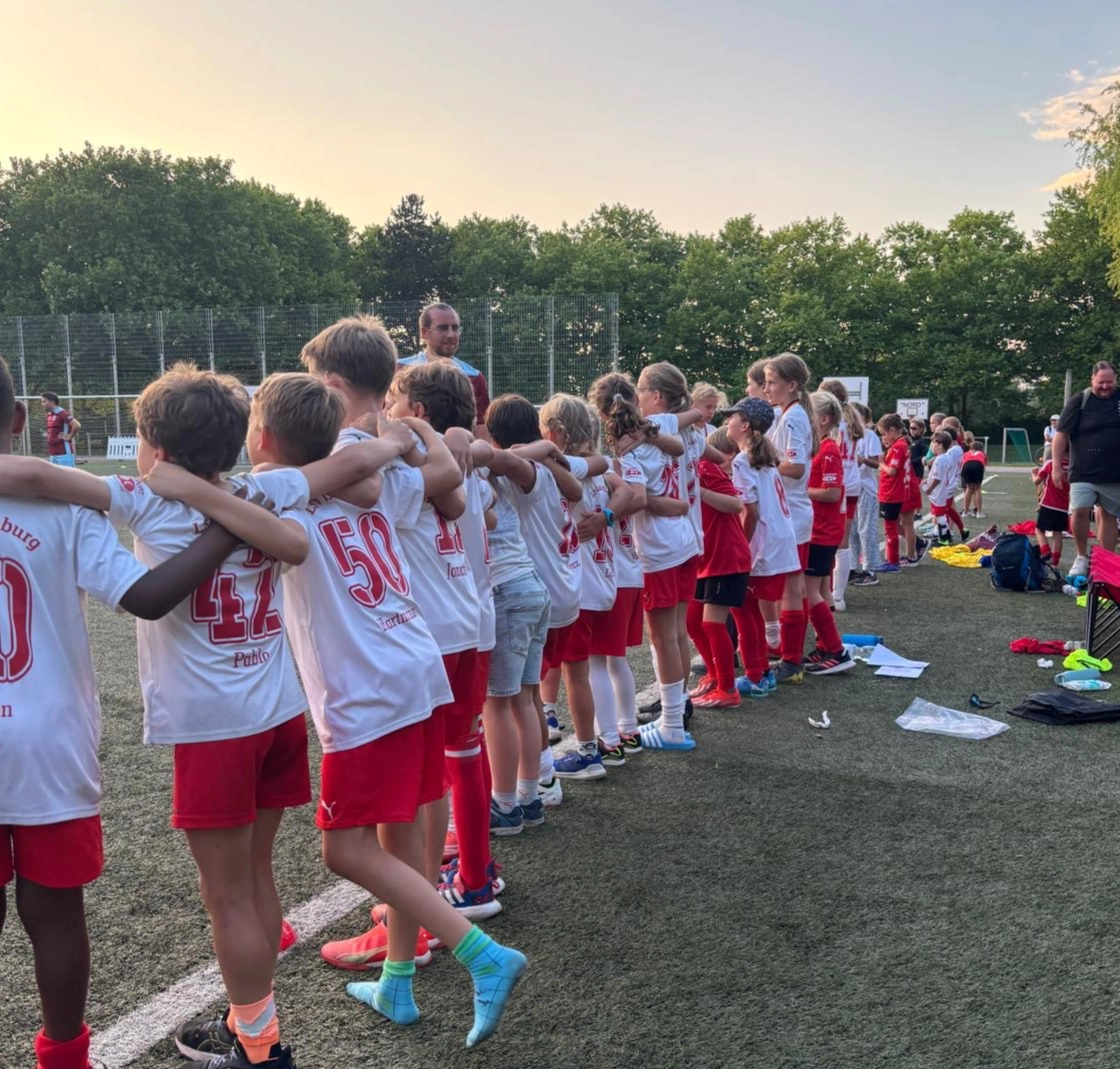 Gruppe von Kindern in weißen und roten Fußballtrikots steht Arm in Arm in einer Reihe auf einem Kunstrasenplatz.