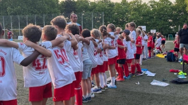 Gruppe von Kindern in weißen und roten Fußballtrikots steht Arm in Arm in einer Reihe auf einem Kunstrasenplatz.