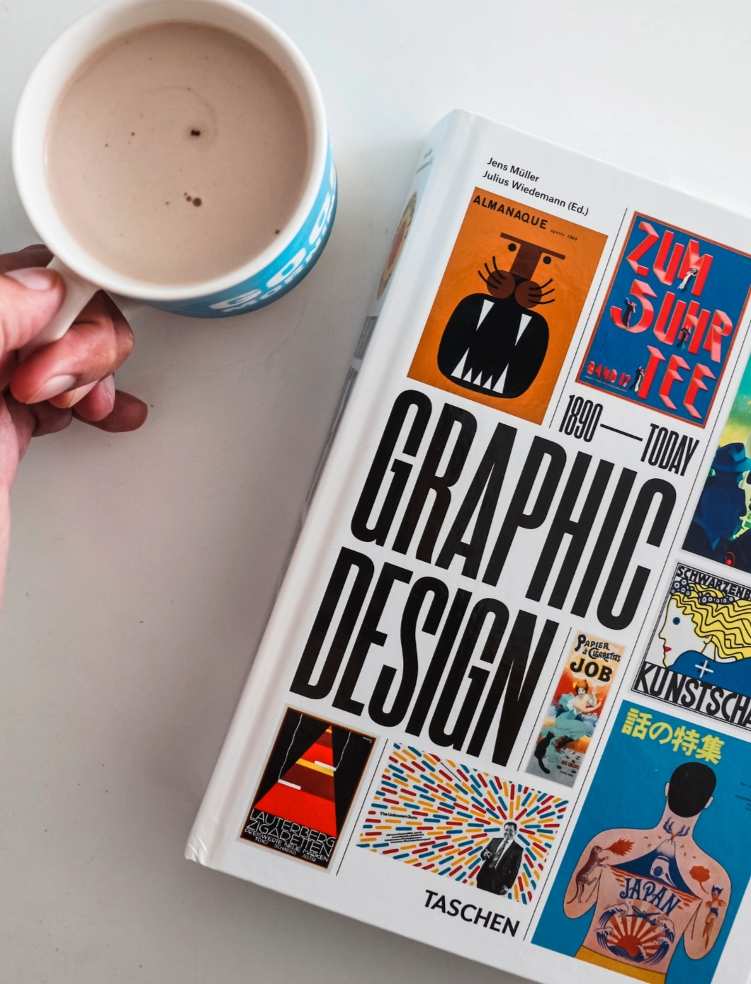 Hand hält eine Tasse Kaffee oder Kakao neben dem Buch „Graphic Design“ von Taschen auf weißem Untergrund.