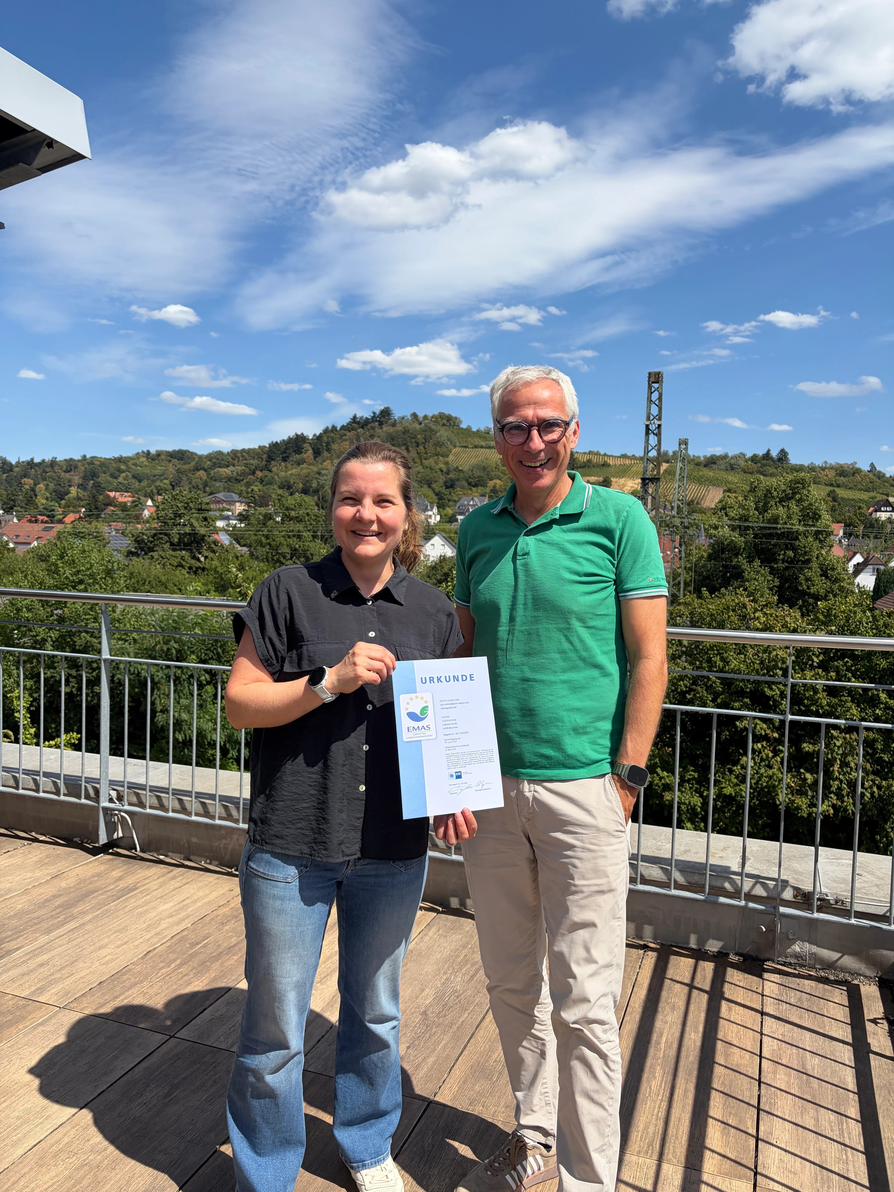 Zwei Personen auf einer Terrasse halten gemeinsam eine Urkunde, im Hintergrund Hügel, Bäume und Wohnhäuser unter blauem Himmel.