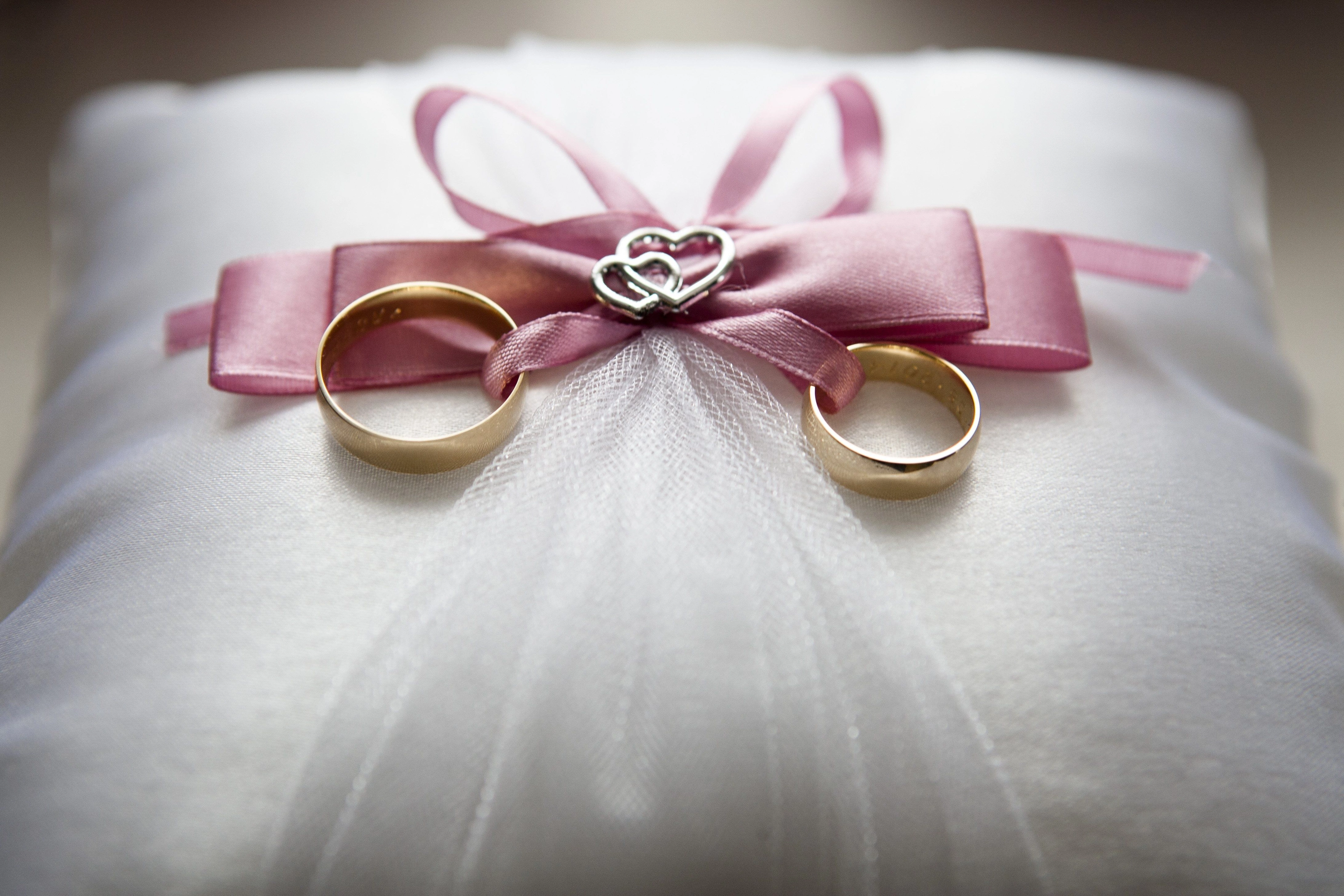 Zwei goldene Ringe auf weißem Ringkissen mit rosa Schleife und kleinem Herzornament.