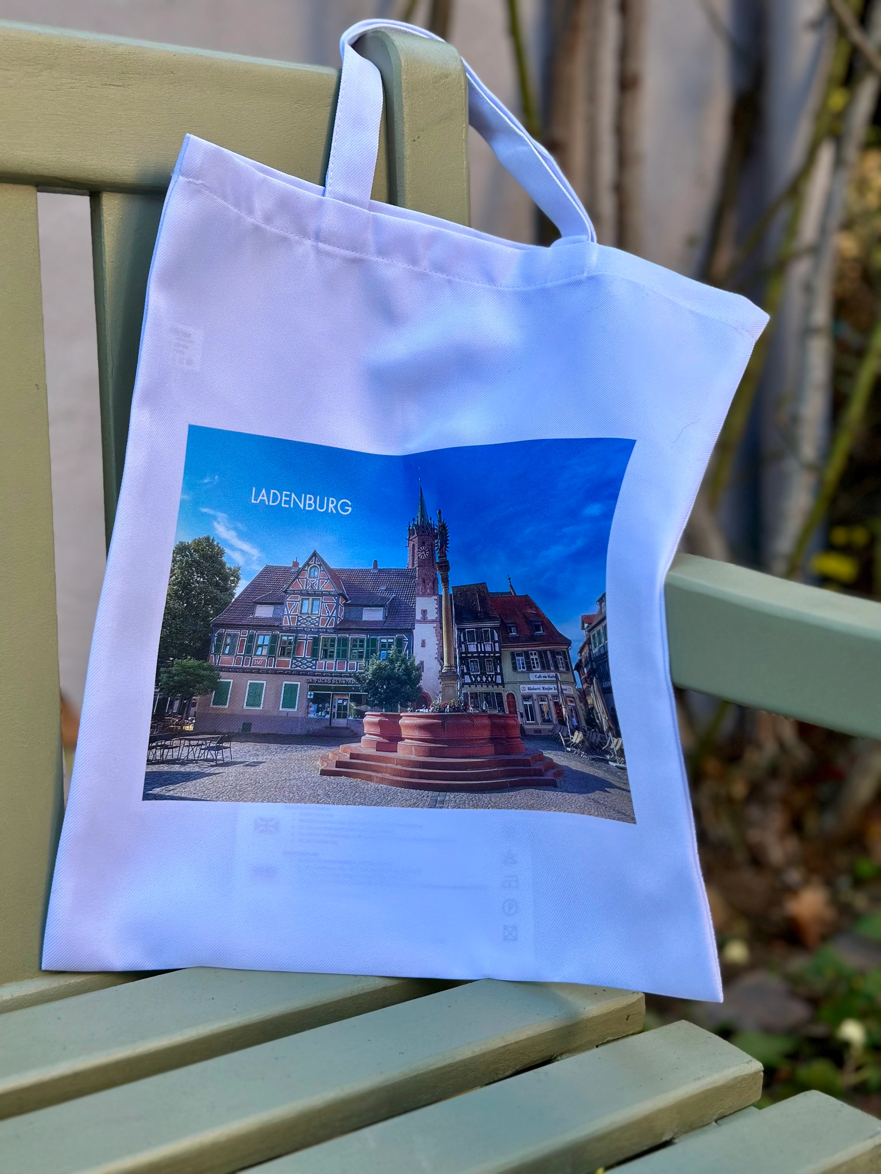 Weiße Stofftasche mit aufgedrucktem Foto eines Platzes und Brunnen in Ladenburg, hängt an einer grünen Gartenbank.