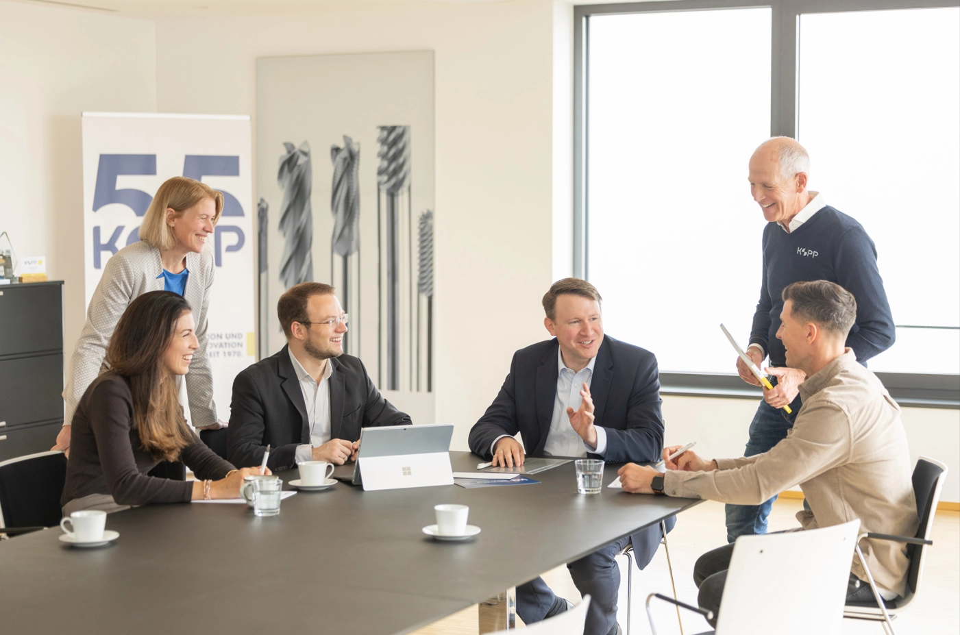 Sechs Erwachsene sitzen und stehen um einen Konferenztisch mit Laptop, Tassen und Papieren und führen eine Besprechung.