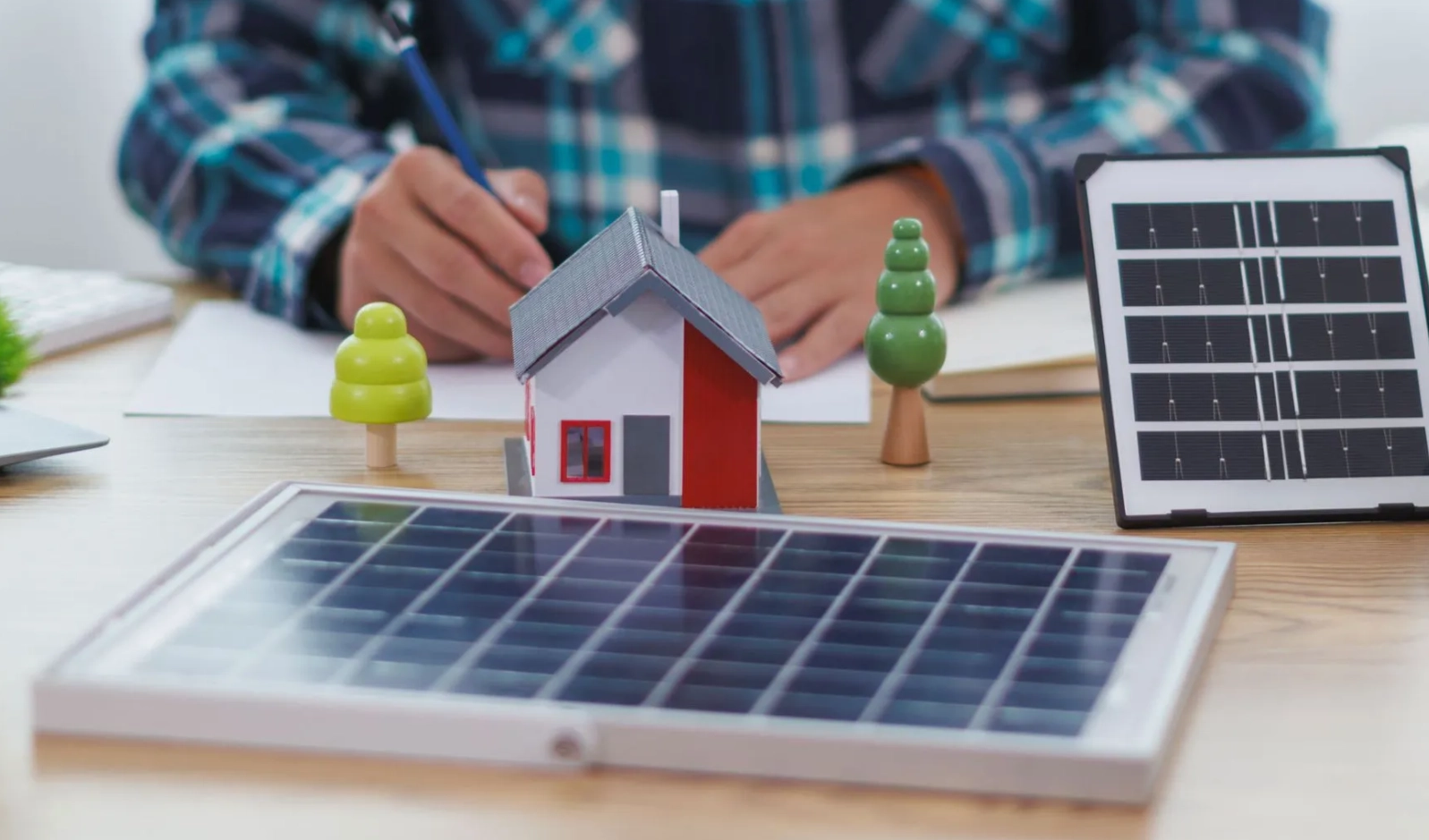 Modellhaus auf einem Tisch mit Solarmodulen und zwei kleinen Bäumen, im Hintergrund eine Person, die schreibt.