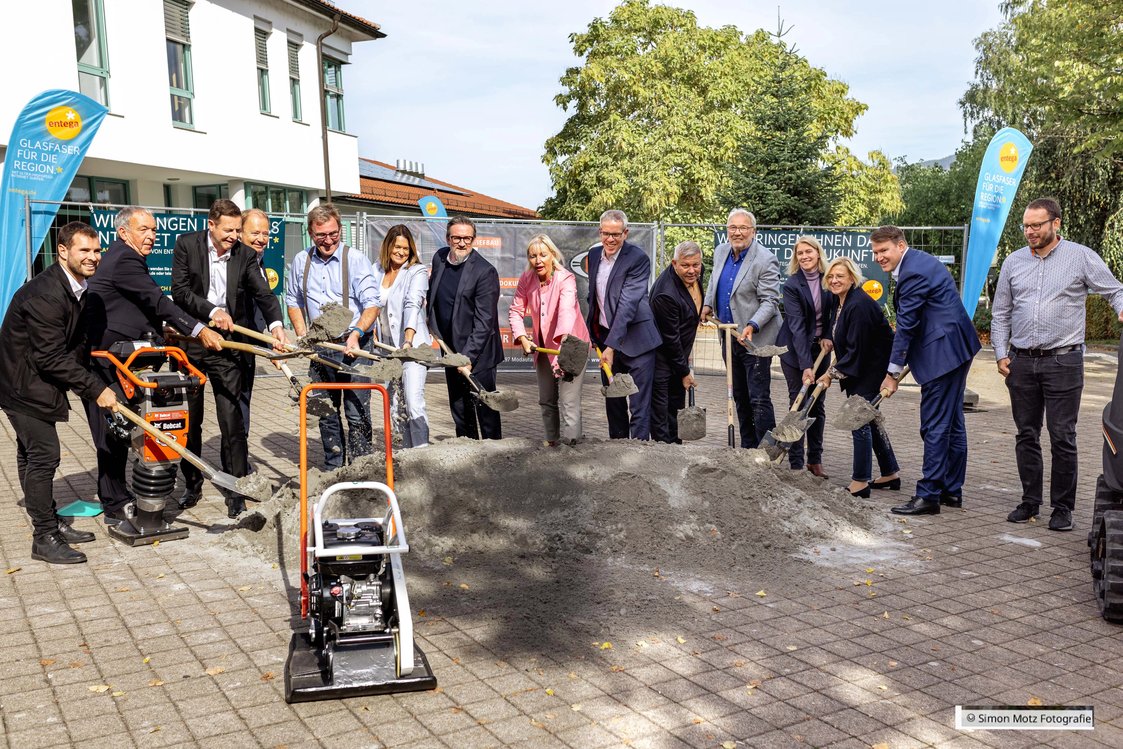 Mehrere Erwachsene in einer Reihe bei einem symbolischen Spatenstich, Schaufeln in einem Erdhaufen; Baustellengeräte im Vordergrund.