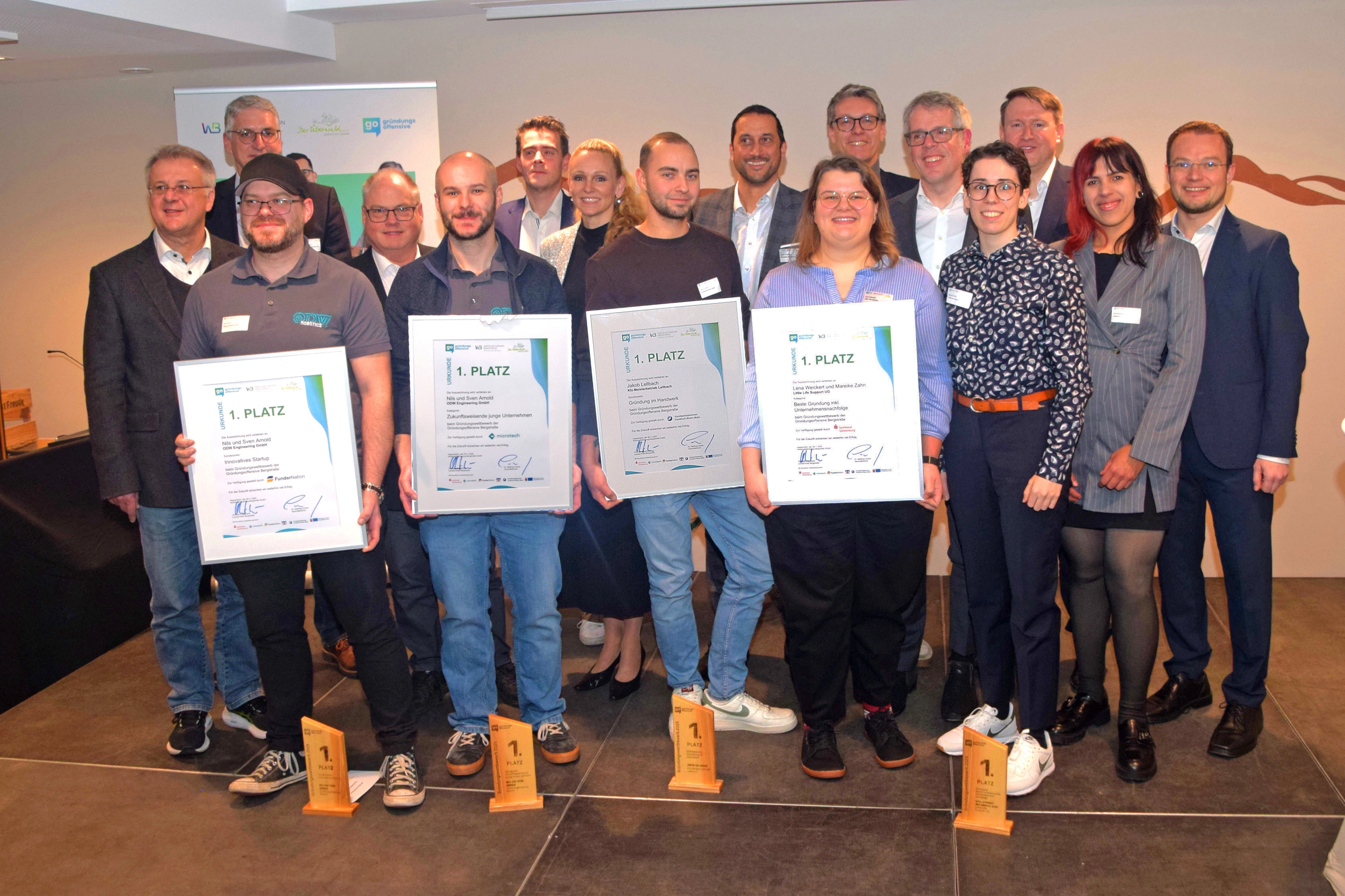 Gruppe von etwa 18 Erwachsenen bei einer Preisverleihung; vier Personen vorne halten gerahmte Urkunden und vor ihnen stehen Holzpokale.