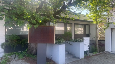 Eingangsbereich eines Gebäudes mit Metall-Schild auf einer Säule, Treppe, Briefkasten und einem großen Baum davor.