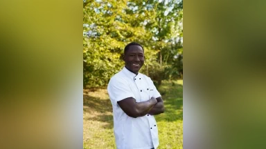 Lächelnder Mann in weißer Kochjacke steht im Freien mit verschränkten Armen.