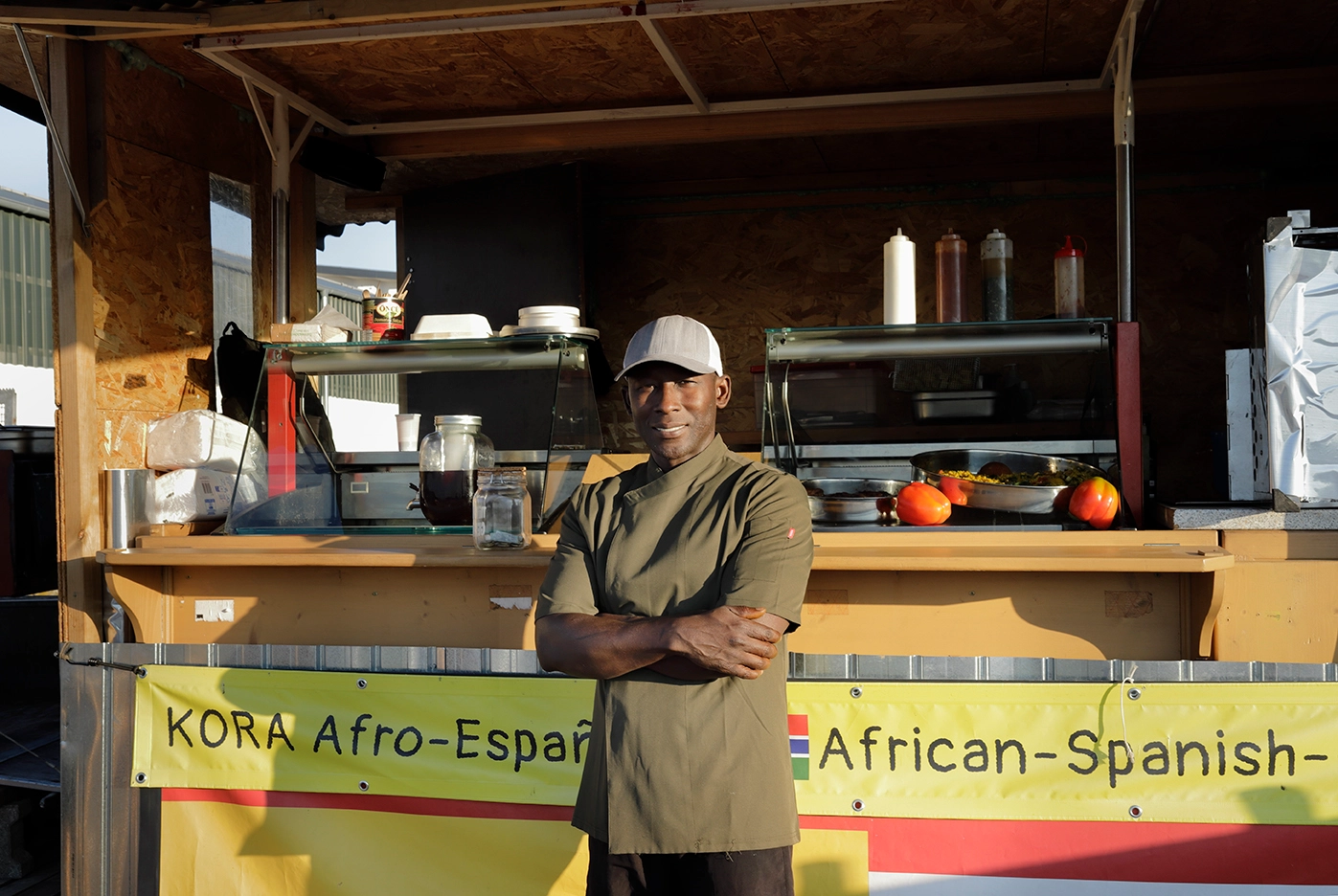 Mann in Kochjacke und Mütze steht mit verschränkten Armen vor einem afrikanisch-spanischen Imbissstand.