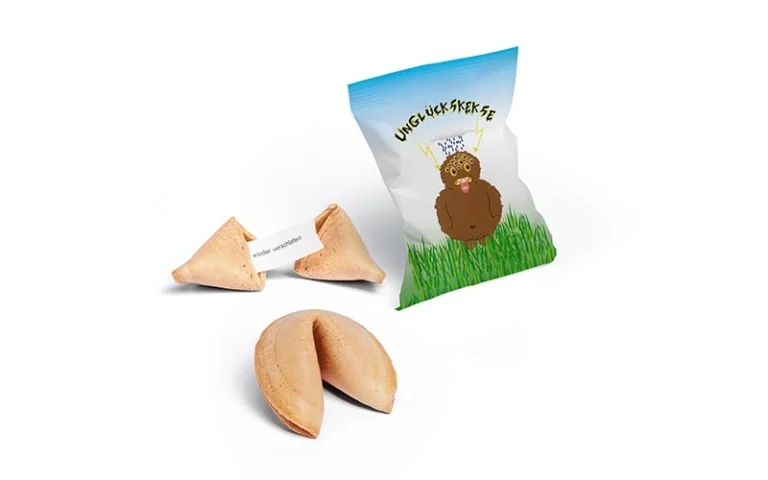 Mehrere Glückskekse, einer geöffnet mit einem Zettel, neben einer Verpackung mit Cartoon und der Aufschrift ‚Unglückskekse‘.