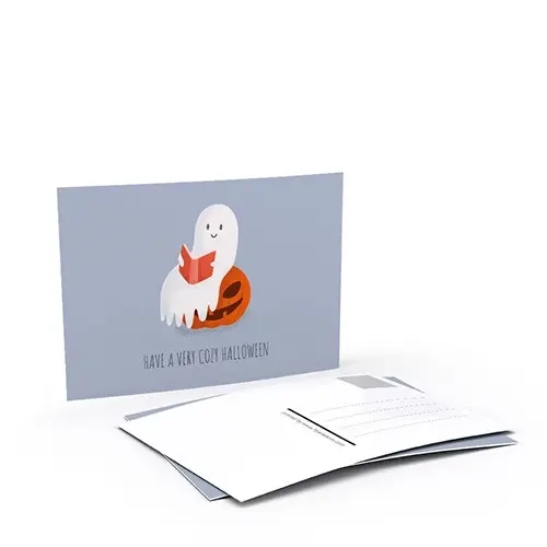 Postkarte mit einem lächelnden Geist, der ein Buch liest und auf einem Kürbis sitzt; Text: ‚Have a very cozy Halloween‘.