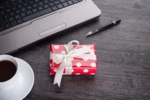 Schreibtisch mit Laptop, Stift, Kaffeetasse und rot-weiß gepunktetem Geschenk mit weißer Schleife.