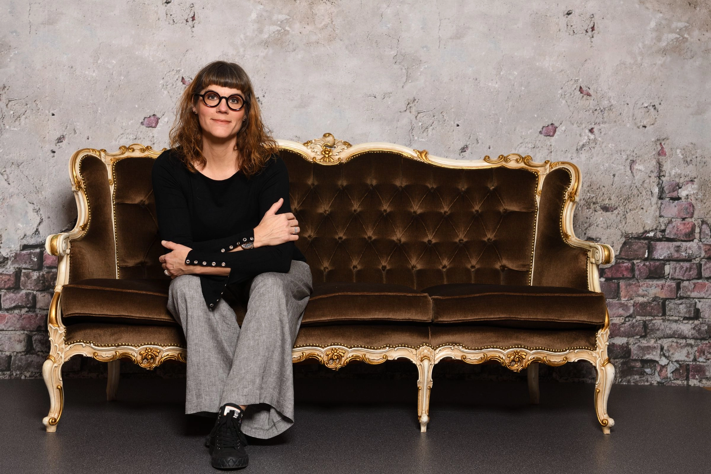 Erwachsene Frau mit Brille sitzt auf einem braunen, barocken Sofa vor einer beschädigten Putz- und Backsteinwand.