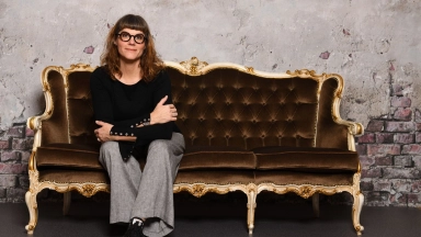 Erwachsene Frau mit Brille sitzt auf einem braunen, barocken Sofa vor einer beschädigten Putz- und Backsteinwand.