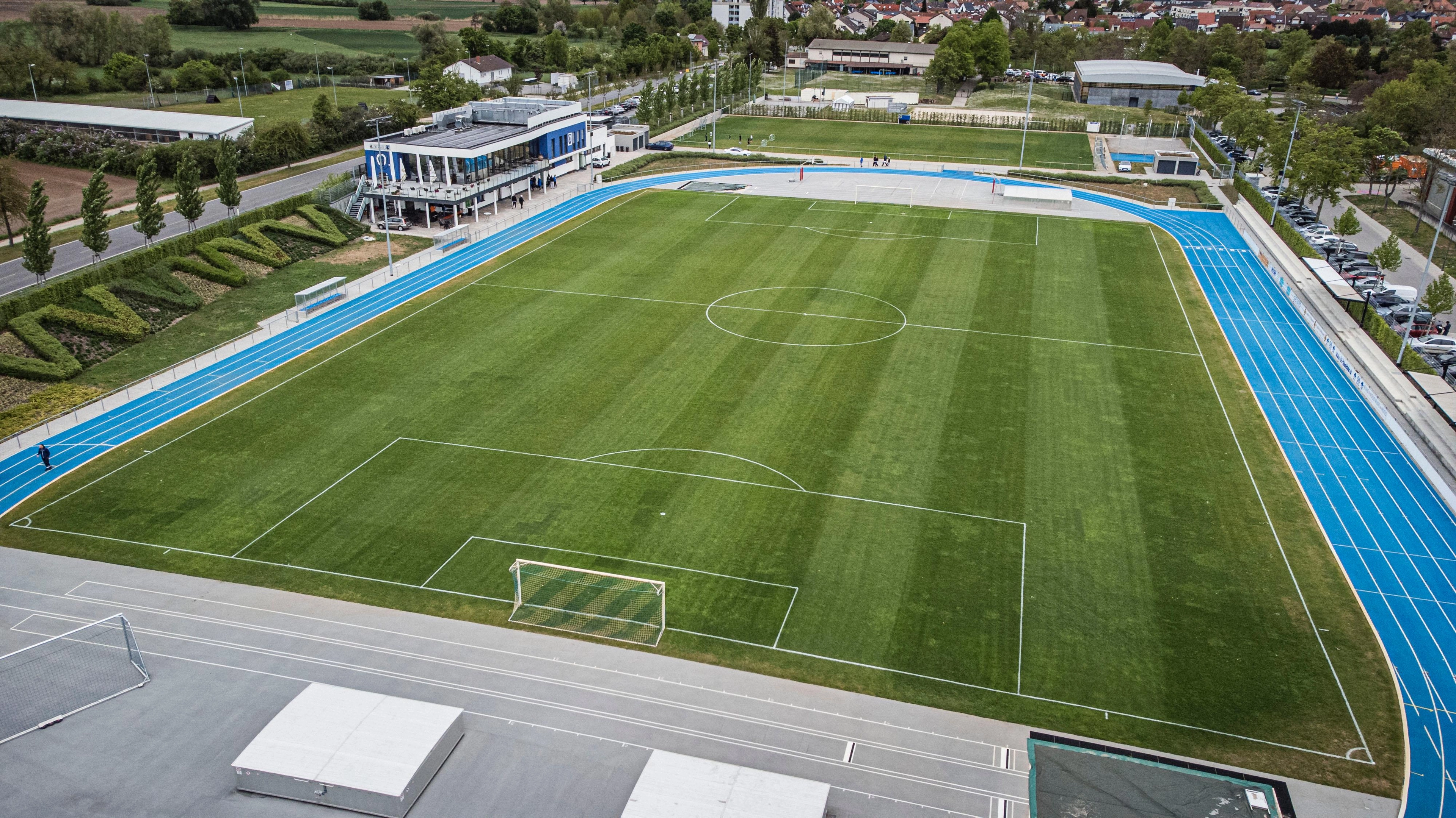 Luftaufnahme eines Fußballrasens mit blauer Leichtathletikbahn und Vereinsgebäude im Hintergrund.