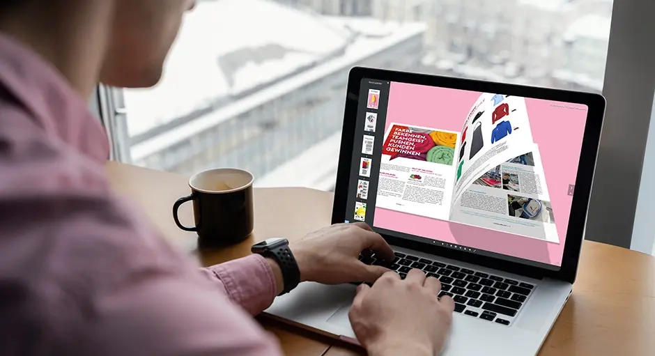 Person an einem Laptop am Fensterplatz, auf dem Bildschirm ein digitales Magazin-Layout, Kaffeetasse auf dem Tisch.