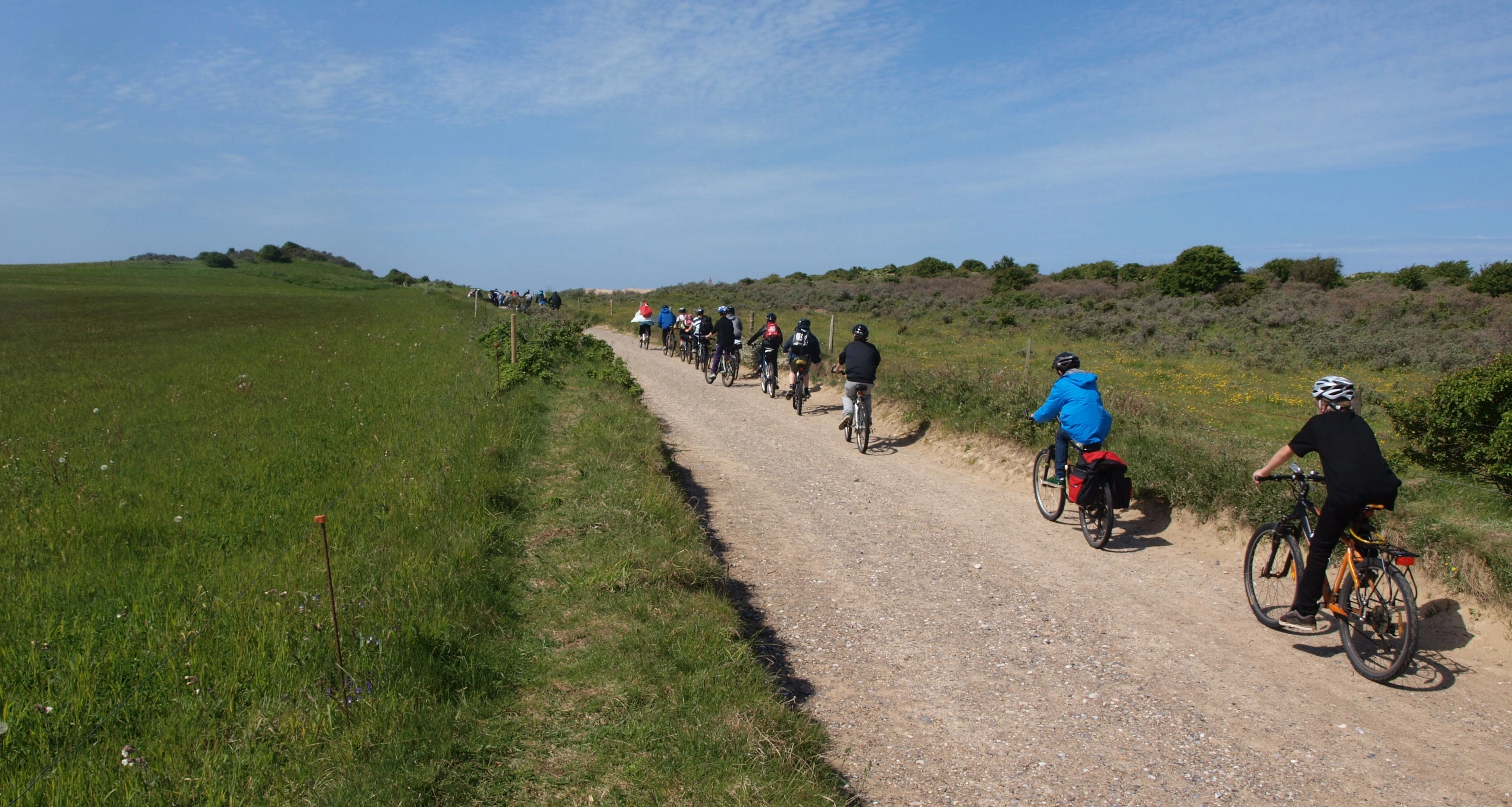 Gruppe von Radfahrern auf einem Schotterweg zwischen grünen Feldern unter blauem Himmel.