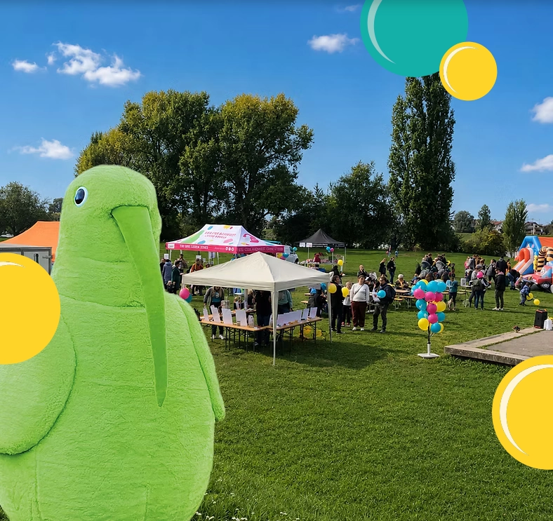 Grünes Vogelförmiges Maskottchen im Vordergrund eines Parkfestes mit Zelten, Besuchern und bunten Luftballons.