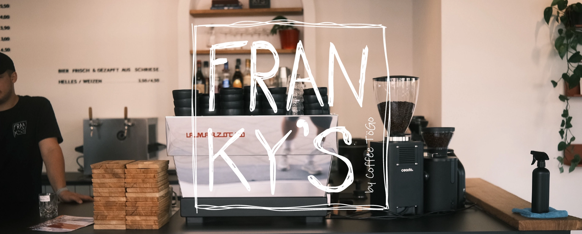 Kaffeetheke mit Espressomaschine, Kaffeemühle, Holzstapeln und einem Mitarbeiter im Hintergrund.