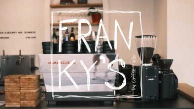Kaffeetheke mit Espressomaschine, Kaffeemühle, Holzstapeln und einem Mitarbeiter im Hintergrund.