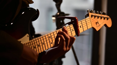 Nahaufnahme einer Hand, die auf dem Hals einer Fender-E-Gitarre mit Kapodaster Akkord greift.
