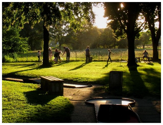 Sonnenbeschienener Park mit Bäumen, mehreren Personen (Erwachsene und Kinder) und Minigolfbahnen im Vordergrund.