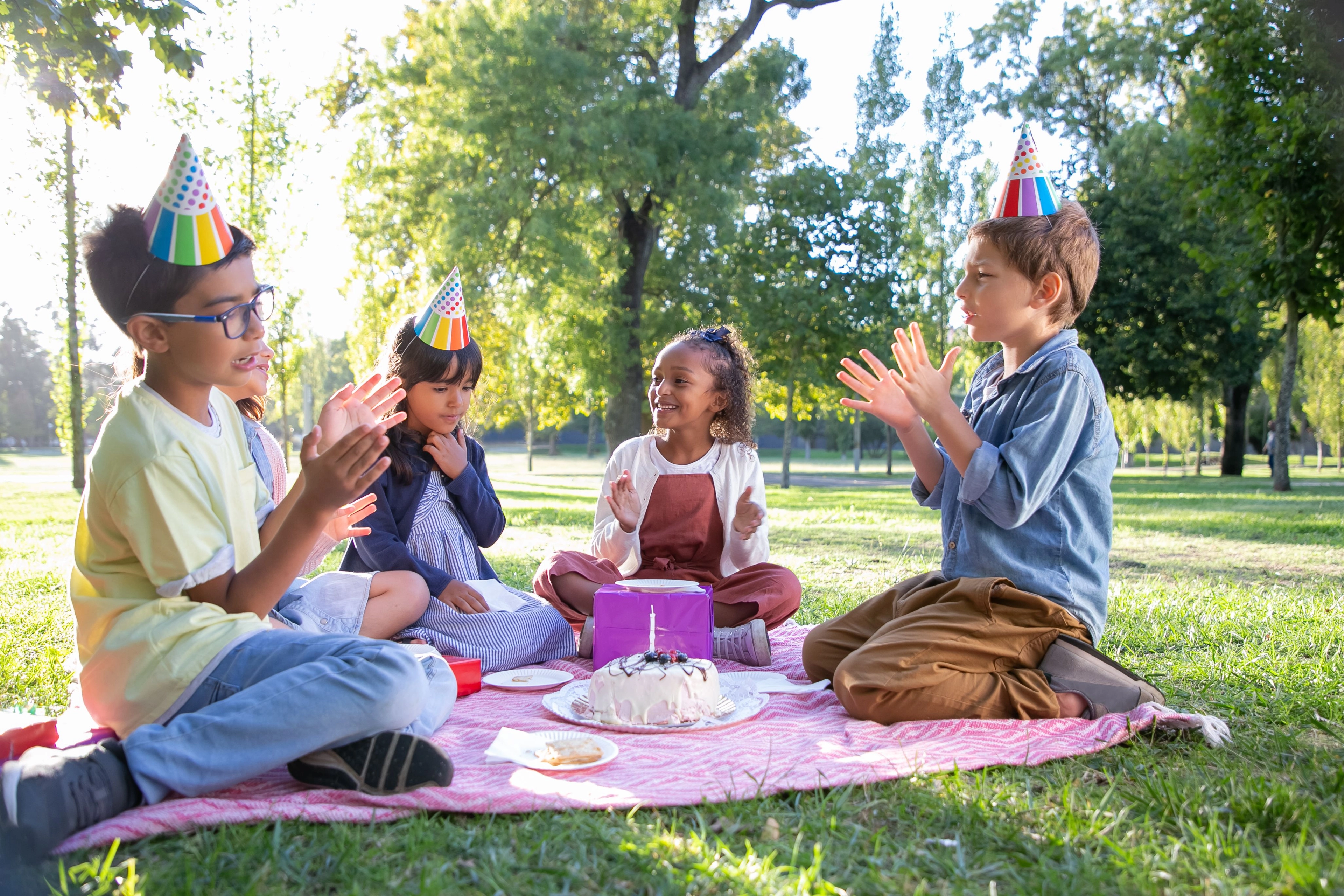 Vier Kinder sitzen auf einer Picknickdecke im Park, tragen Partyhüte und klatschen um einen Geburtstagskuchen mit Kerze.