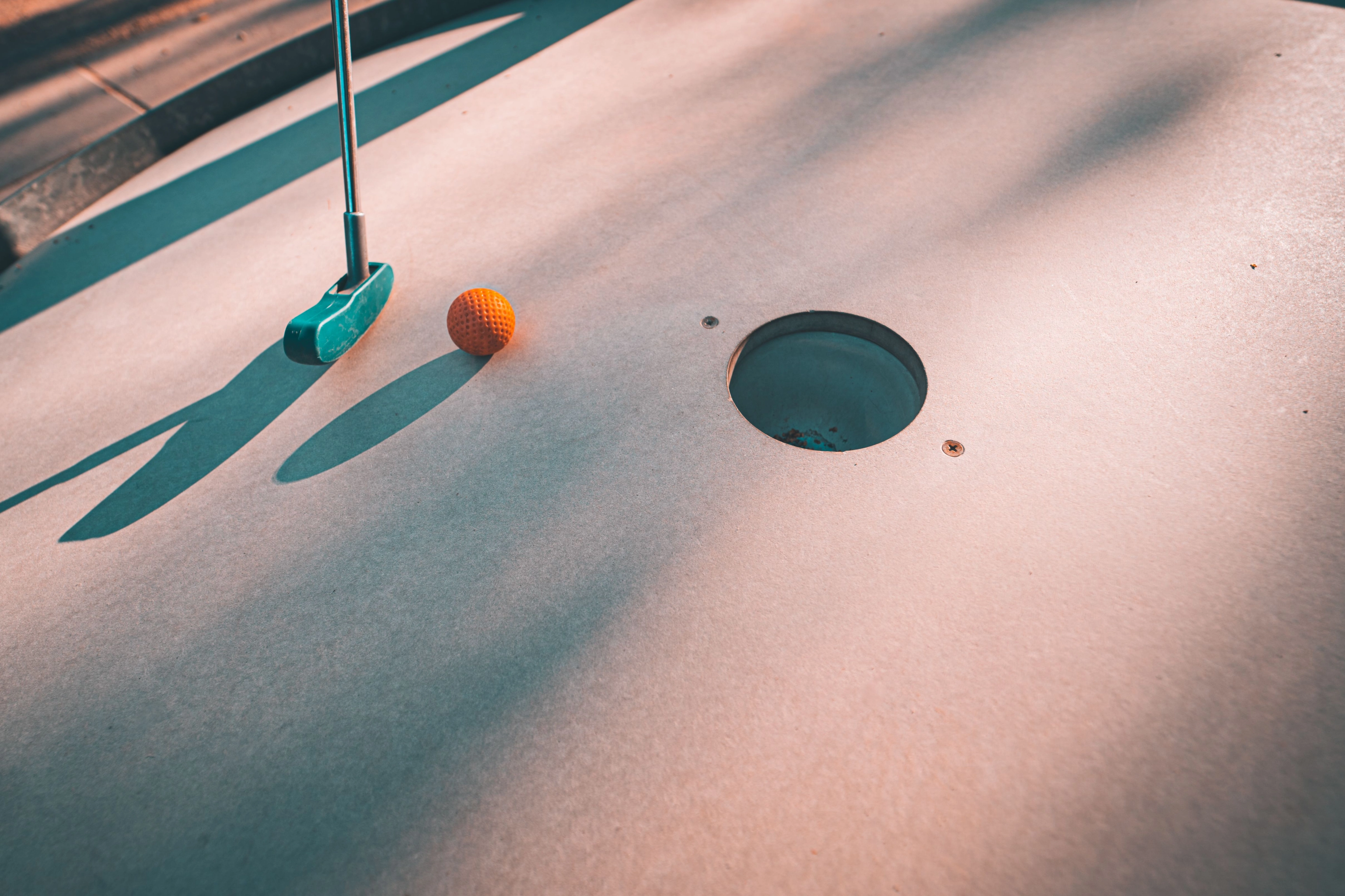 Nahaufnahme eines Minigolf-Putters und eines orangefarbenen Golfballs neben einem Loch auf der Spieloberfläche.