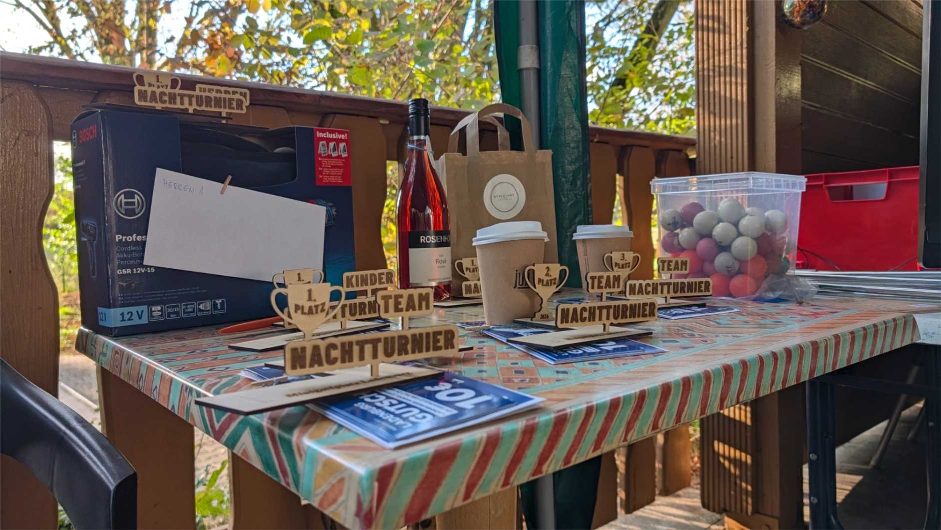 Tisch auf einer Terrasse mit mehreren Preis-Trophäen mit Schriftzug 'Nachtturnier', Flasche, Pappbechern, einer Plastikbox mit Bällen und weiteren Verpackungen.