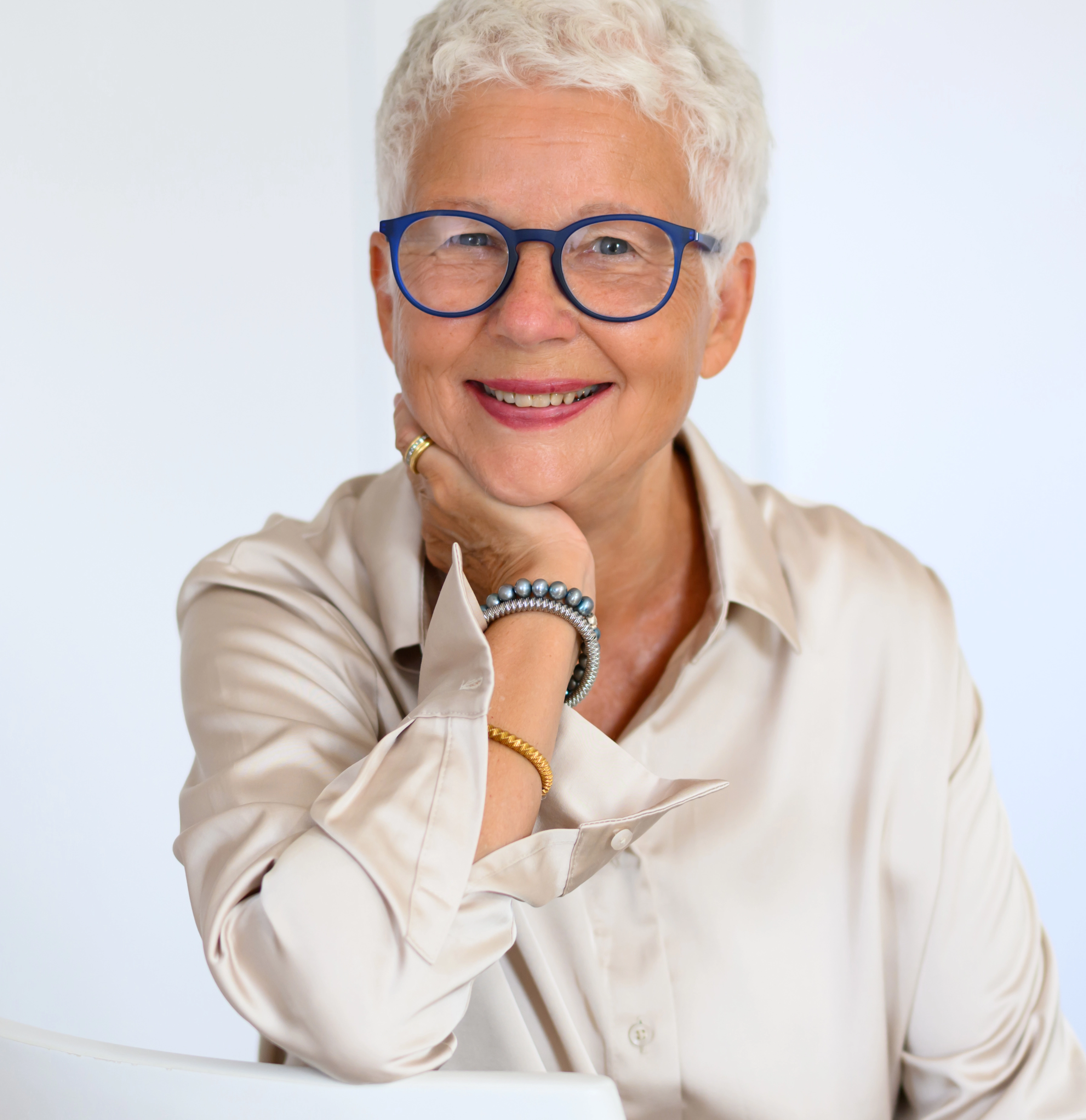 Porträt einer älteren lächelnden Frau mit kurzen weißen Haaren und blauer Brille, sitzend und die Hand am Kinn.